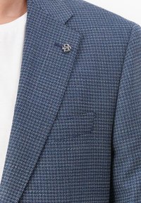 Mans blazer i mörkblå strukturerad tyg med ett rutigt mönster, med en silverfärgad detaljer på slaget. Vit t-shirt med rund hals synlig.