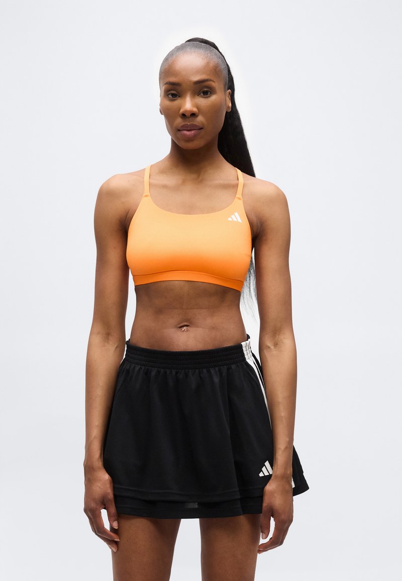 Αθλητική γυναίκα με μακριά μαλλιά, φορώντας πορτοκαλί αθλητικό σουτιέν και μαύρη φούστα Adidas, στέκεται μπροστά σε λευκό φόντο, κοιτάζοντας προς τα εμπρός.