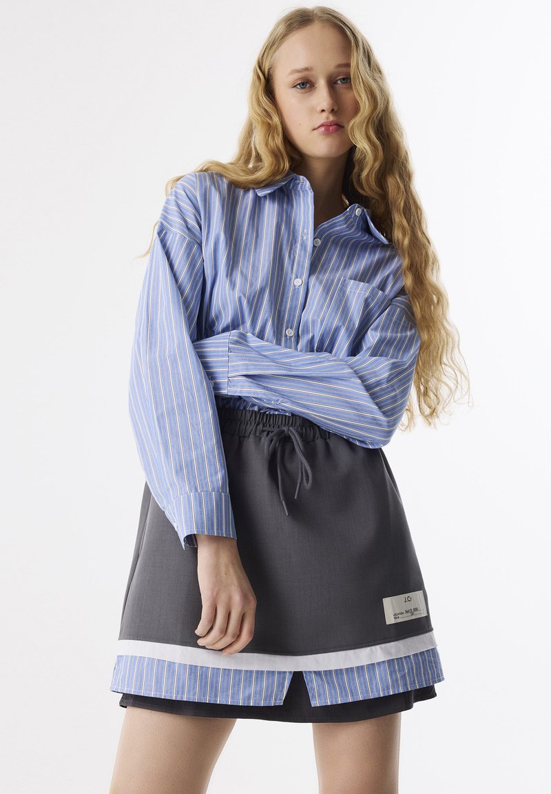 Chemise à rayures bleues et blanches à boutons, associée à une jupe grise avec une taille élastique et des accents blancs en couches au niveau de l'ourlet.