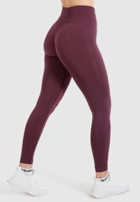 Bordeaux-hinnoiset legginsit, joissa on sileä ja joustava pinta. Niissä on sivusauma, jossa on hillitty brändäys, ja ne on yhdistetty valkoisiin urheilusandaaleihin.
