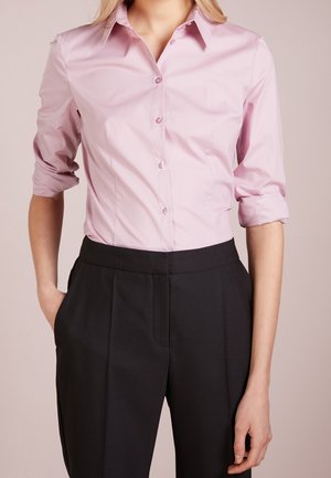 Persona che indossa una camicia rosa chiaro con bottoni e maniche arrotolate e pantaloni neri a vita alta, con le mani parzialmente nelle tasche.