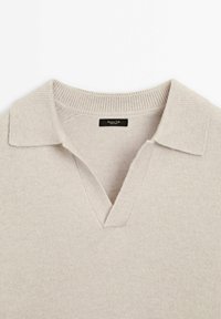 Pull en tricot beige avec col en V et col chemise. Texture lisse, col côtelé, et étiquette de marque minimaliste à l'intérieur.