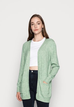 Cardigan à manches longues en tricot vert clair, avec un devant ouvert, deux poches avant et des poignets côtelés. Porté par-dessus un crop top blanc.