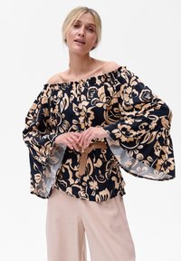 Bluzka bez ramiączek w kolorze granatowym z dużym beżowym nadrukiem kwiatowym, z bufiastymi rękawami i falbanką na dekolcie, zestawiona z beżowym paskiem i jasnymi spodniami.