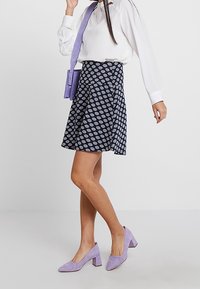 Falda navy con un patrón geométrico, combinada con una blusa blanca y zapatos de tacón cuadrado en color lavanda. Bolso morado adicional llevado en bandolera.