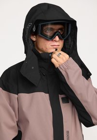 Snowboardjack met een zwart en lichtroze kleurblokontwerp, duurzame stof, verstelbare capuchon en logodetail op de borst.