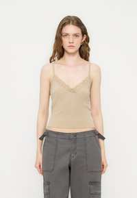 Γυναίκα με κυματιστά μαλλιά που φοράει μπεζ καμιζόλ με δαντέλα και γκρι παντελόνια cargo, στέκεται μπροστά σε απλό ανοιχτό φόντο.