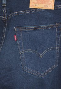 Mörka denimjeans med slim fit, kontrasterande gula sömmar och en läderlapp med märke och storleksdetaljer på midjebandet.