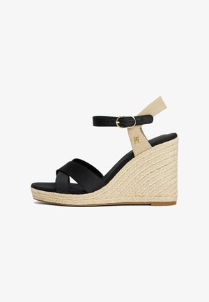 Sandales compensées en cuir noir avec des brides entrecroisées, une bride de cheville beige et un talon en jute tissé. Un détail en boucle dorée ajoute une touche subtile.