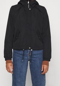 Svart zip-up jacka med tryckknappslappar och dragsko i nederkant, bärs över bruna långärmade tröjor och blå jeans.