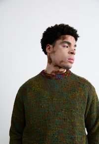 Knitted sweater with a ribbed texture, primarily green, featuring colorful specks of red, blue, and yellow. Crew neck design.