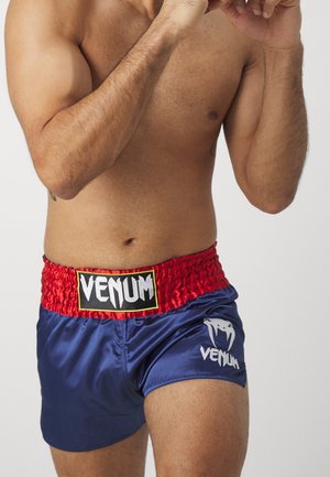 Mężczyzna sportowiec w niebieskich i czerwonych spodenkach muay thai Venum z podniesionymi rękami w pozycji bokserskiej na jednolitym tle.