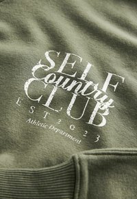 Žalias megztinis, pagamintas iš minkštos medžiagos, su baltu nusidėvėjusiu užrašu "SELF Country CLUB EST 2023" ir "Sporto departamentas."