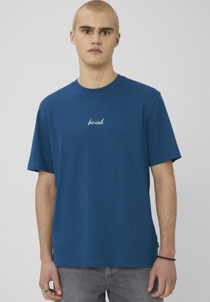 Jeune homme aux cheveux blonds courts portant un t-shirt bleu avec le mot « break » sur la poitrine et un jean gris, debout devant un fond neutre.