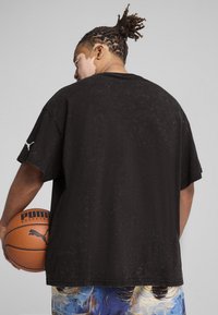Sort oversized T-shirt med korte ærmer, der har en tekstureret overflade og et hvidt Puma-logo på venstre ærme. Basketball holdes under armen.