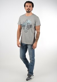 Siva majica kratkih rukava s plavim grafičkim tekstom koji kaže "SURF CLUB." Uparena s svijetloplavim trapericama i sivim tenisicama s bijelim detaljima.