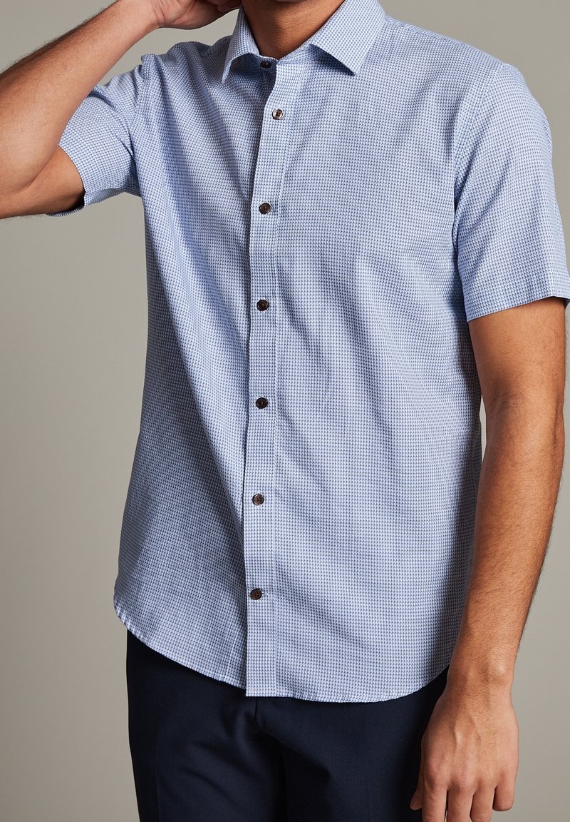 Homme portant une chemise bleu clair à manches courtes avec un petit motif à carreaux et un pantalon foncé, main touchant le cou.