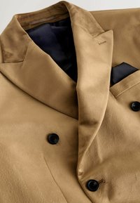 Svetlohnedý dvojradový kabát vyrobený z hladkej látky s čiernymi gombíkmi a kontrastným námorníckym vreckovým štvrtkom. Pozoruhodné jemné textúry a klopy.