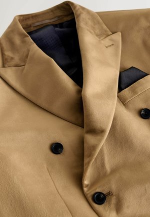 Kamelihalliga kaksiknööbiga jakk, valmistatud siledast kangast, mustade nööpide ja kontrastse tumesinise taskurätiku ning märkimisväärsete pehmete tekstuuride ja kraedega.