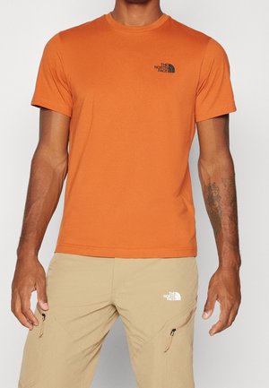 Mann trägt ein orangefarbenes Kurzarm-T-Shirt und beige Cargohosen, beide mit The North Face-Logos, vor einem einfarbigen Hintergrund.