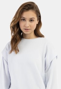 Světle modrá mikina s uvolněným střihem, která má spadlé ramenní švy a hladkou texturu. Minimalistický design s širokým výstřihem.