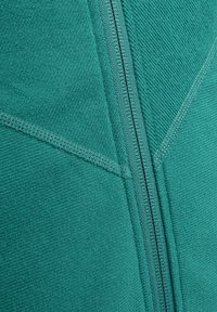 Tealfarbener Hoodie mit Reißverschluss aus einem strukturierten, fleeceähnlichen Material, das sich durch kontrastierende Nähte und einen schrägen Nahtdesign auszeichnet.