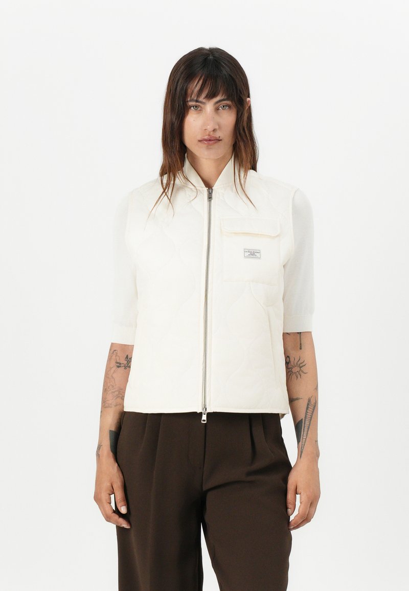 Gilet trapuntato avorio con colletto alto, maniche corte, zip frontale e tasca sul petto con piccolo etichetta logo. Pantaloni marroni sotto.