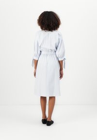 Mulher com cabelo encaracolado a usar um vestido de comprimento até ao joelho, listrado em azul claro e branco, com mangas atadas, de costas, em fundo branco.