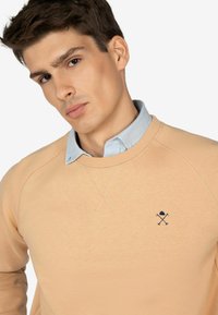Licht perzik sweatshirt met een ronde hals, voorzien van een klein marineblauw geborduurd logo. Gelaagd over een lichtblauw overhemd met kraag.