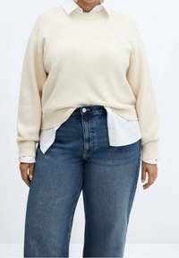 Pull en tricot beige avec une texture côtelée porté sur une chemise blanche à col. Assorti à un jean large taille haute bleu foncé.