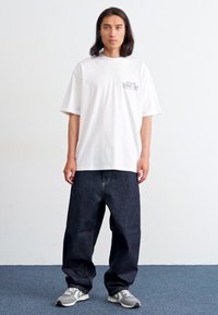 Hvit t-skjorte med kort erme og trykt logo på brystet, med mørkeblå baggy jeans og grå joggesko. Minimalistisk design.