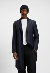 Cappotto lungo blu navy con vestibilità sartoriale, revers tagliati e sei bottoni neri, abbinato a una camicia bianca e pantaloni neri. Berretto a costine blu navy.