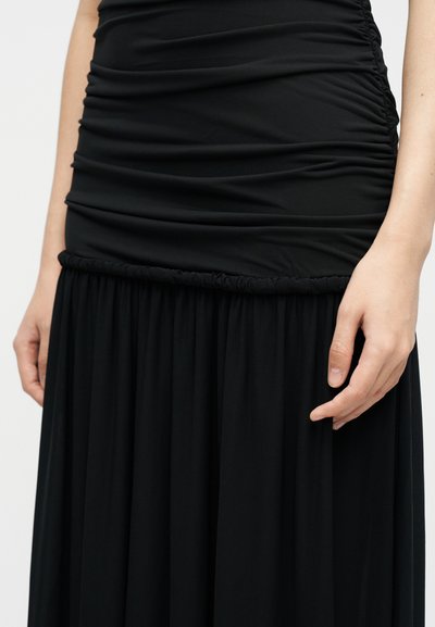 Κοντινό πλάνο ενός ατόμου που φοράει μαύρη κορυφή με ζάρες και μαύρη ροή φούστα, με το ένα χέρι χαλαρό στο πλάι του.