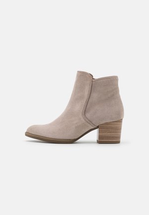 Beige suède enkelboot met een gestapelde houten blokhak en een zijrits, in profiel weergegeven op een witte achtergrond.