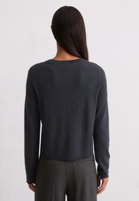 Sieviete ar garām tumšām matēm, tērpta pieguļošā tumši pelēkā garajā piedurknē un augstās vidukļa pelēkās svītrainās biksēs, stāv pretī baltai siena.