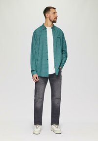 Zielona koszula w kratę na białym t-shircie, w połączeniu z szarymi dżinsami z przetarciami i jasnymi trampkami. Model stoi na jednolitym tle.