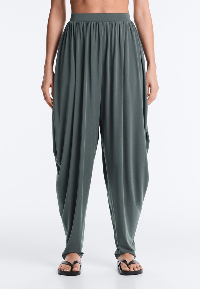 Pantalon large vert foncé fluide avec taille froncée porté par une personne se tenant devant un fond blanc uni.