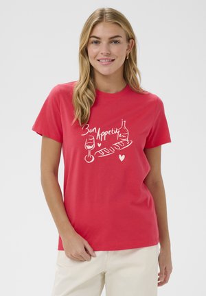 Junge Frau mit langen blonden Haaren, die ein rotes T-Shirt mit weißem "Bon Appetit"-Text sowie Abbildungen eines Weinglases, einer Flasche und eines Baguettes trägt.