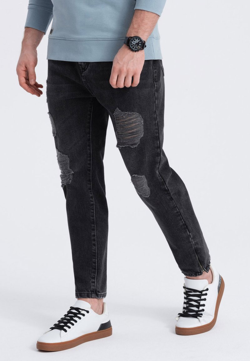 Jeans de mezclilla negros con parches desgastados, ajuste entallado. Combinados con zapatillas blancas con detalles negros y suelas de goma marrón.