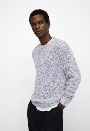 Uomo con capelli corti e ricci che indossa un maglione lavorato a maglia bianco e blu sopra una camicia bianca e pantaloni neri, in piedi con una mano nella tasca.