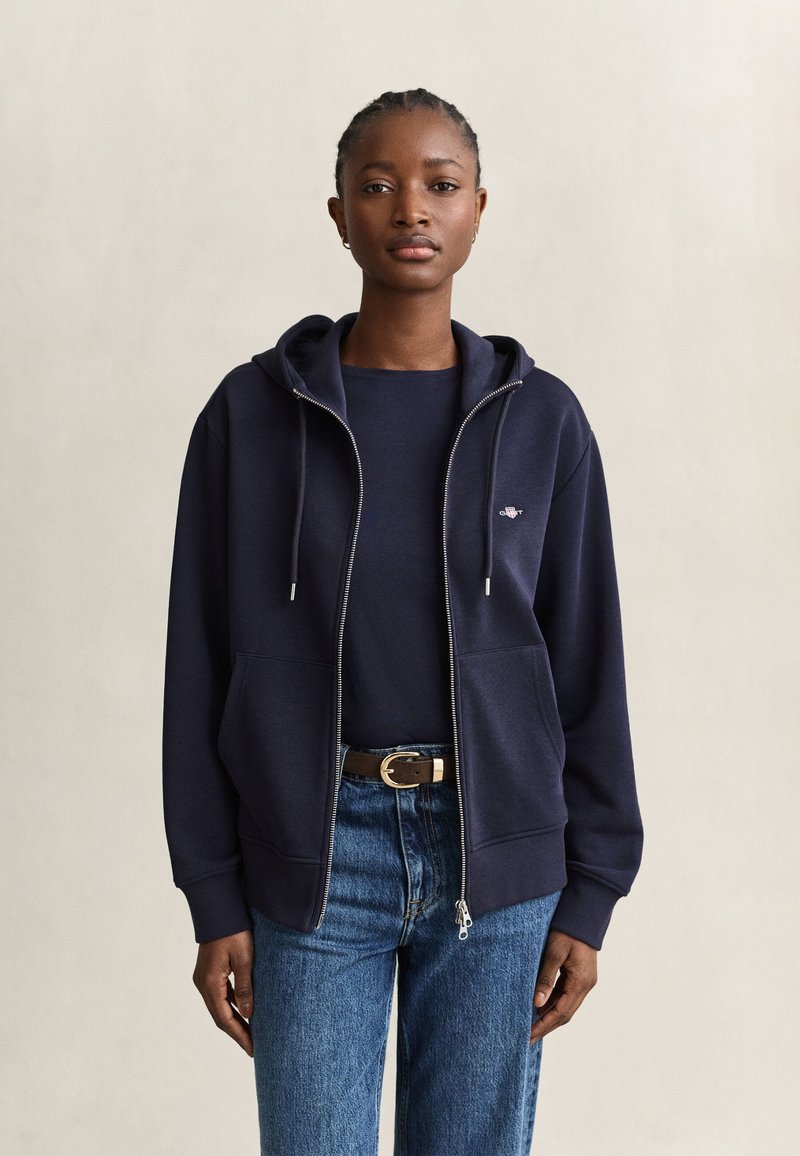 Kvinde iført en mørkeblå lynlåshættetrøje, mørkeblå skjorte, blå jeans med bælte, der står mod en ensartet lys baggrund.