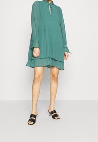 Turkusowa sukienka z lekkiego materiału, z plisami, długim rękawem i wiązaniem przy szyi. Połączona z czarnymi mulesami na obcasie z podwójnymi paskami.