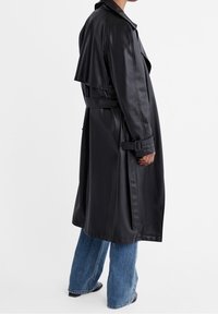 Trench nero in tessuto liscio, con vita cinturata e patta sulla spalla. Abbinato a jeans a gamba larga color azzurro chiaro.