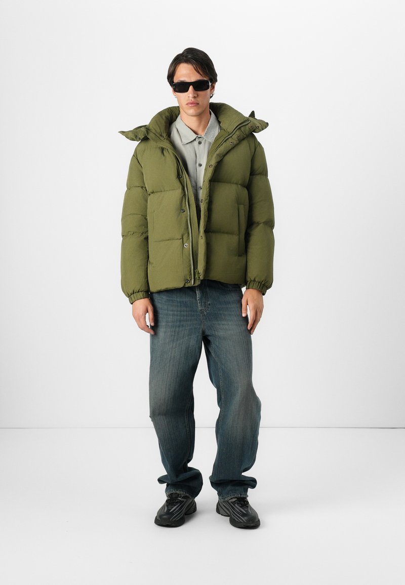 Chaqueta puffer verde con capucha, textura acolchada y puños elásticos, llevada sobre una camisa gris con cuello y unos vaqueros de mezclilla azules. Zapatillas oscuras.