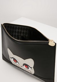 Musta nahkainen pieni laukku, jossa on kohokuvioitu kissan kasvot, joissa on punaisia yksityiskohtia ja kimaltavia koristeita. Kullattu vetoketju ja riipus tuovat näyttäviä metalliosia.