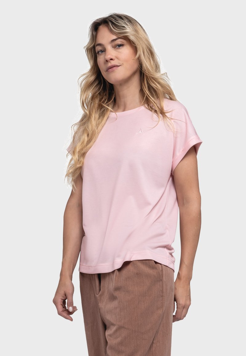 Helles rosa T-Shirt mit kurzen Ärmeln und rundem Ausschnitt sowie einem dezenten Logo auf der Brust; kombiniert mit strukturierten braunen Hosen.