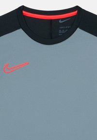 Svetlo siva športna majica z črnim ovratnikom in rameni, rdečim Nike swoosh logotipom na prsih ter rdečim detajlom na notranjem ovratniku.