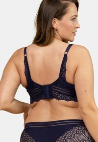 Soutien-gorge en dentelle bleu marine avec bretelles réglables, bordure en dentelle texturée et fermeture à crochets, présentant un dos lisse et des accents décoratifs.