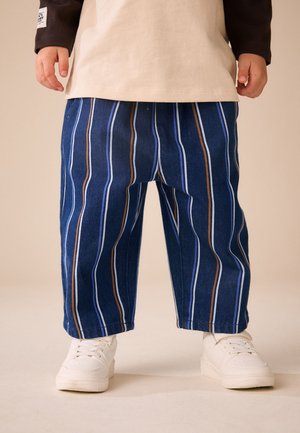 Barn iført blåstripede bukser, hvide sneakers og en beige bluse med lange ærmer, står på en beige gulv.