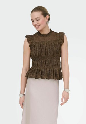 Žena sa usmieva, má na sebe hnedý čipkovaný top bez rukávov s volánmi a svetlo béžovú sukňu, doplnené striebornými kruhovými náušnicami a svetlými náramkami.